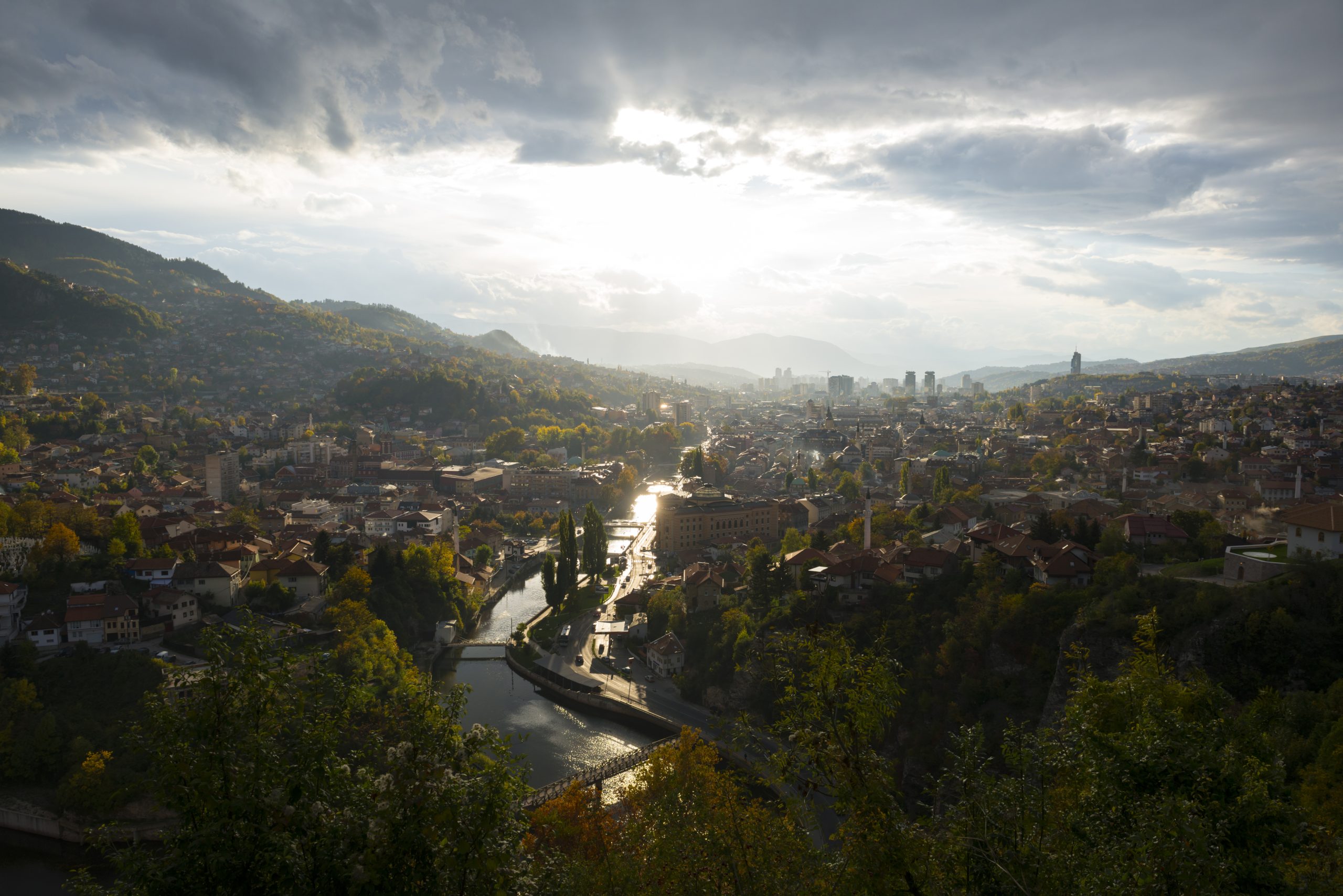 Sarajevo and the Miljacka River seen from a hillside beneath a sunlit cloudy sky